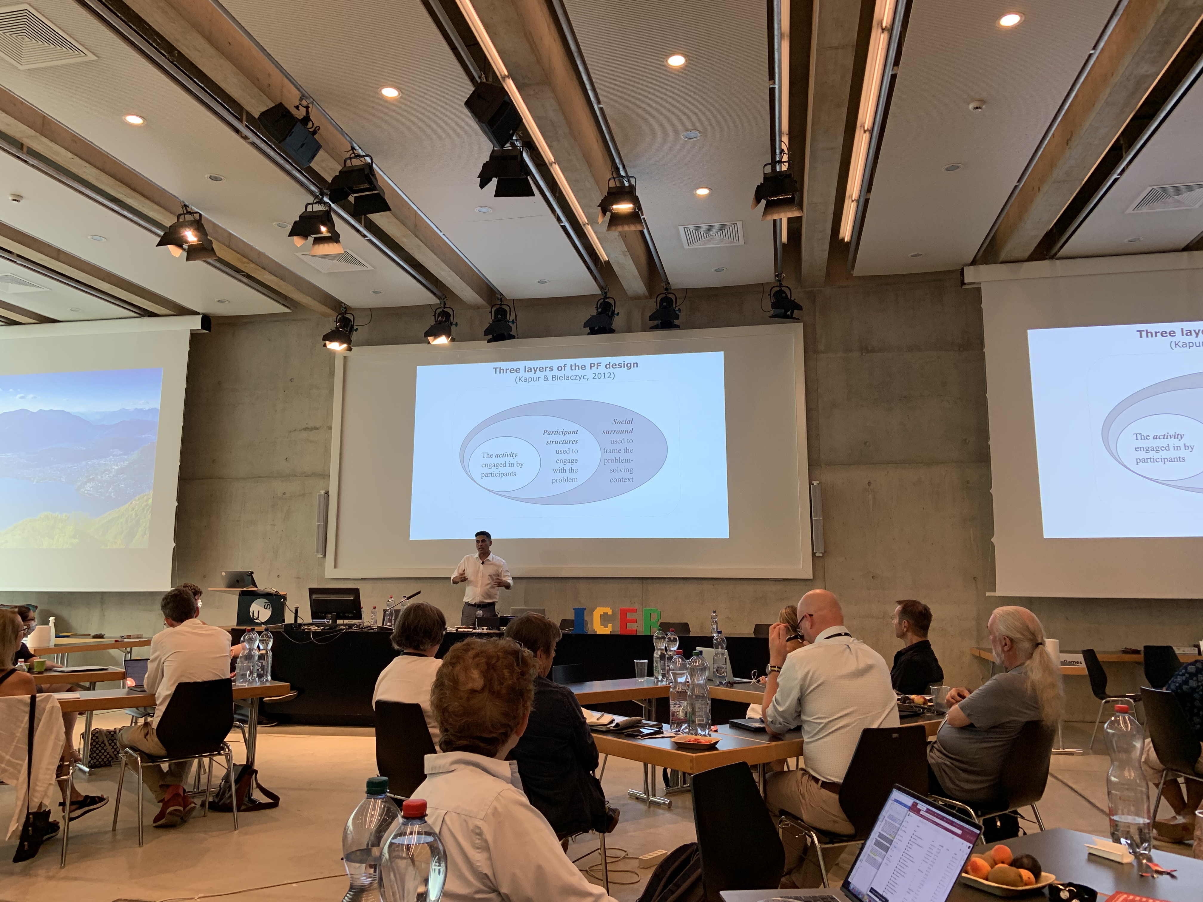 Manu Kapur standing at the ICER stage, presenting the three layers of productive failure design: activity, structures to engage, and social surroundings.