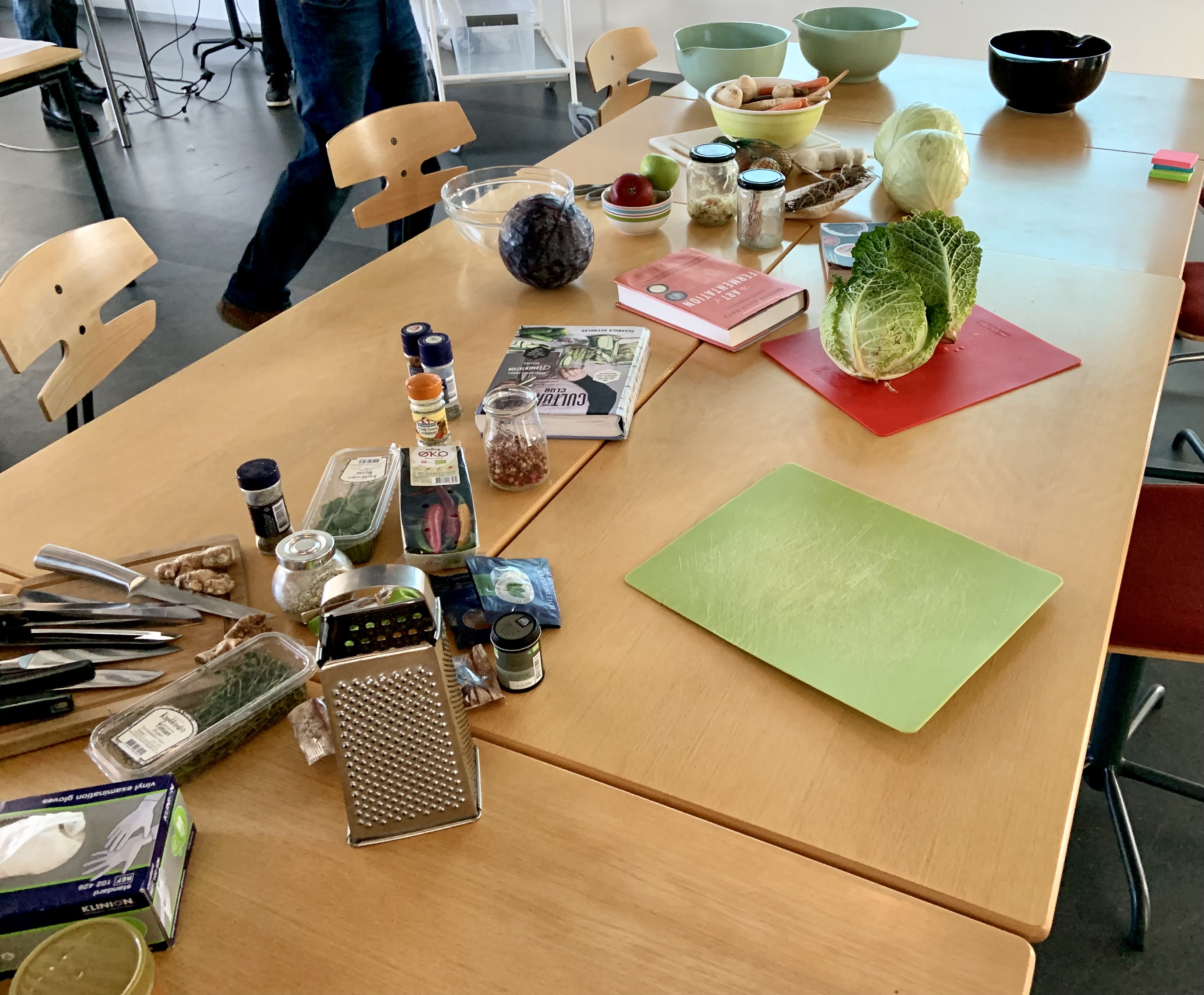 A set of connected tables containing stashes of ingredients and tools for fermenting: vegetables and spices, cookbooks, knives, cuttingboards and a grater.