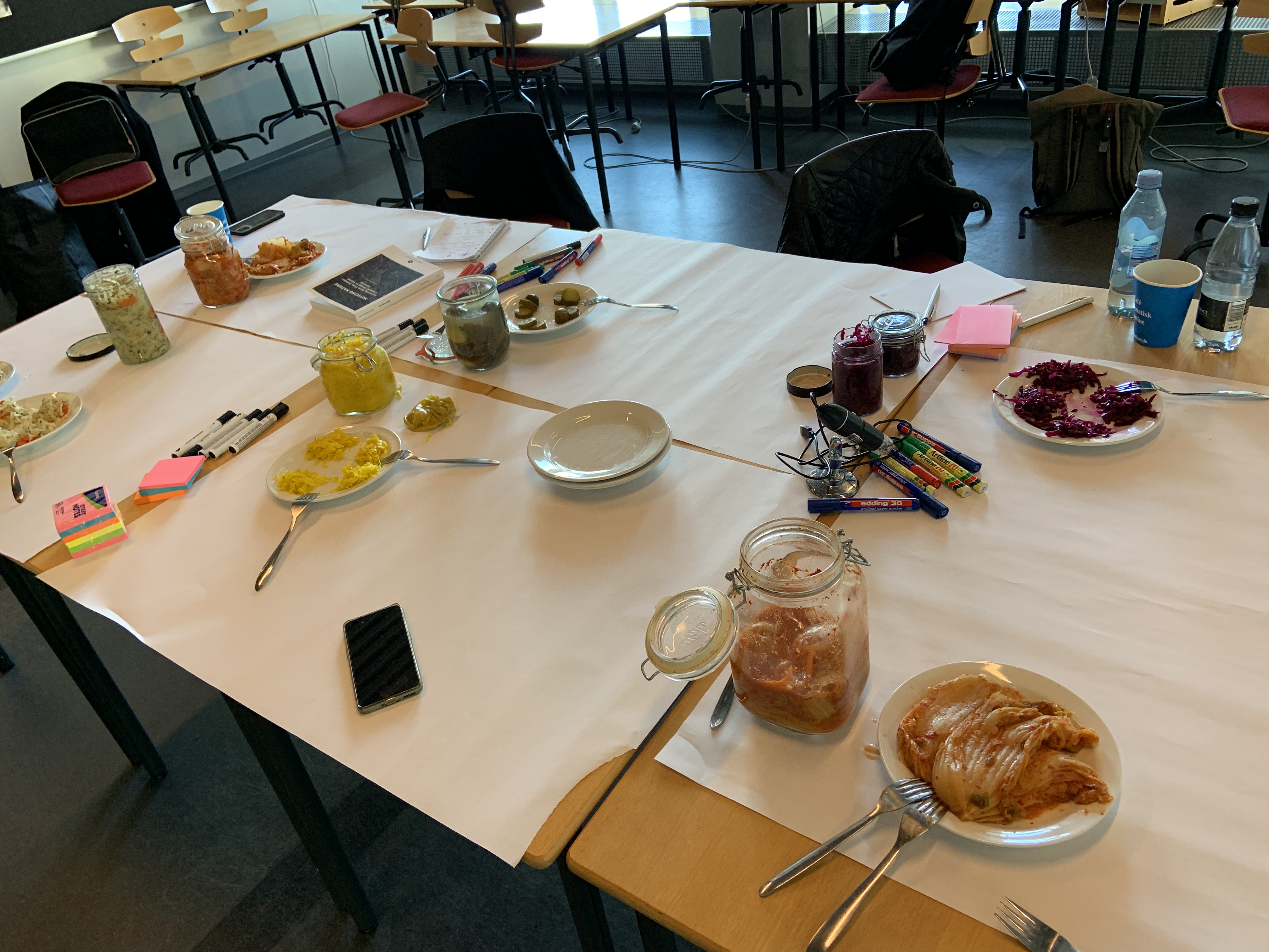 A rectangle of tables covered in large pieces of white paper. On top, there are jars of two month old ferments, some plates, forks and markers.