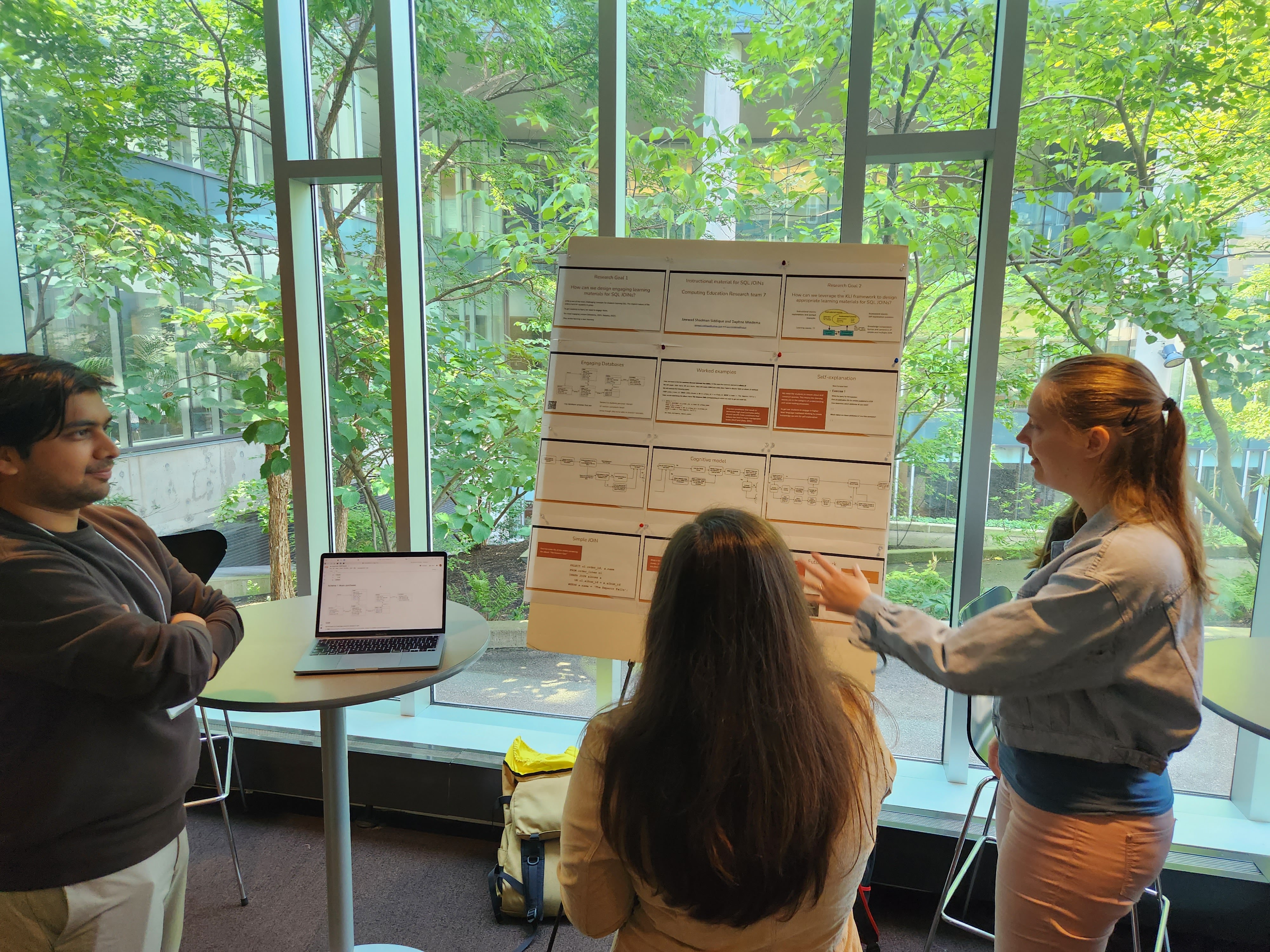 Center in the image is a poster board containing 12 slides turned into a poster. The poster has the title Instructional Material for SQL JOINs. On the right-hand side, Daphne is gesturing toward the poster and explaining some concepts to the person attending, who is standing in the middle of the frame. Jawwad stands to the left, listening.