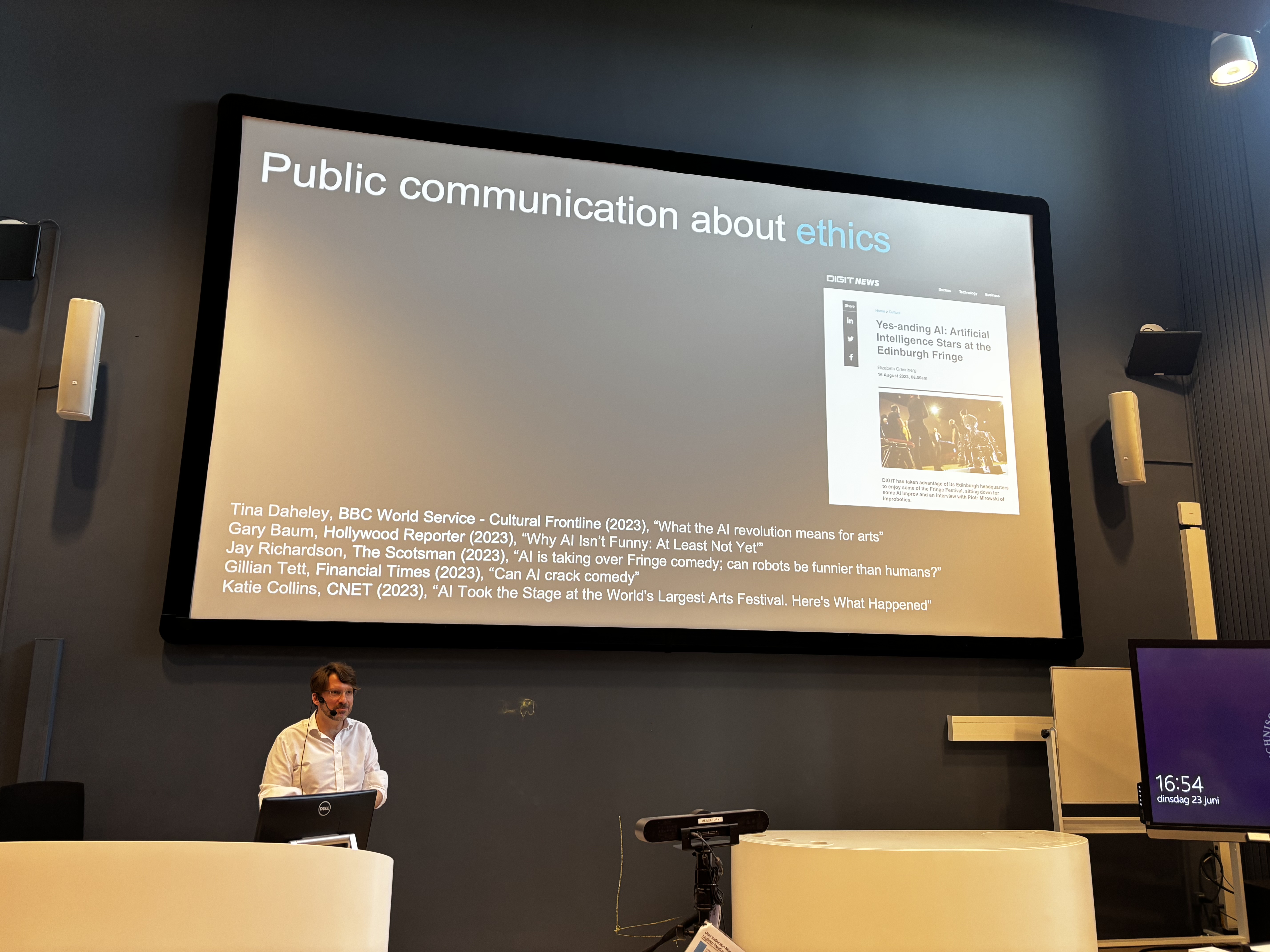 A picture of Piotr giving his talk, in front of a slide containing titles of news articles about their theatre work, including 'What the AI revolution means for arts' and 'Why AI isn't funny: at least not yet