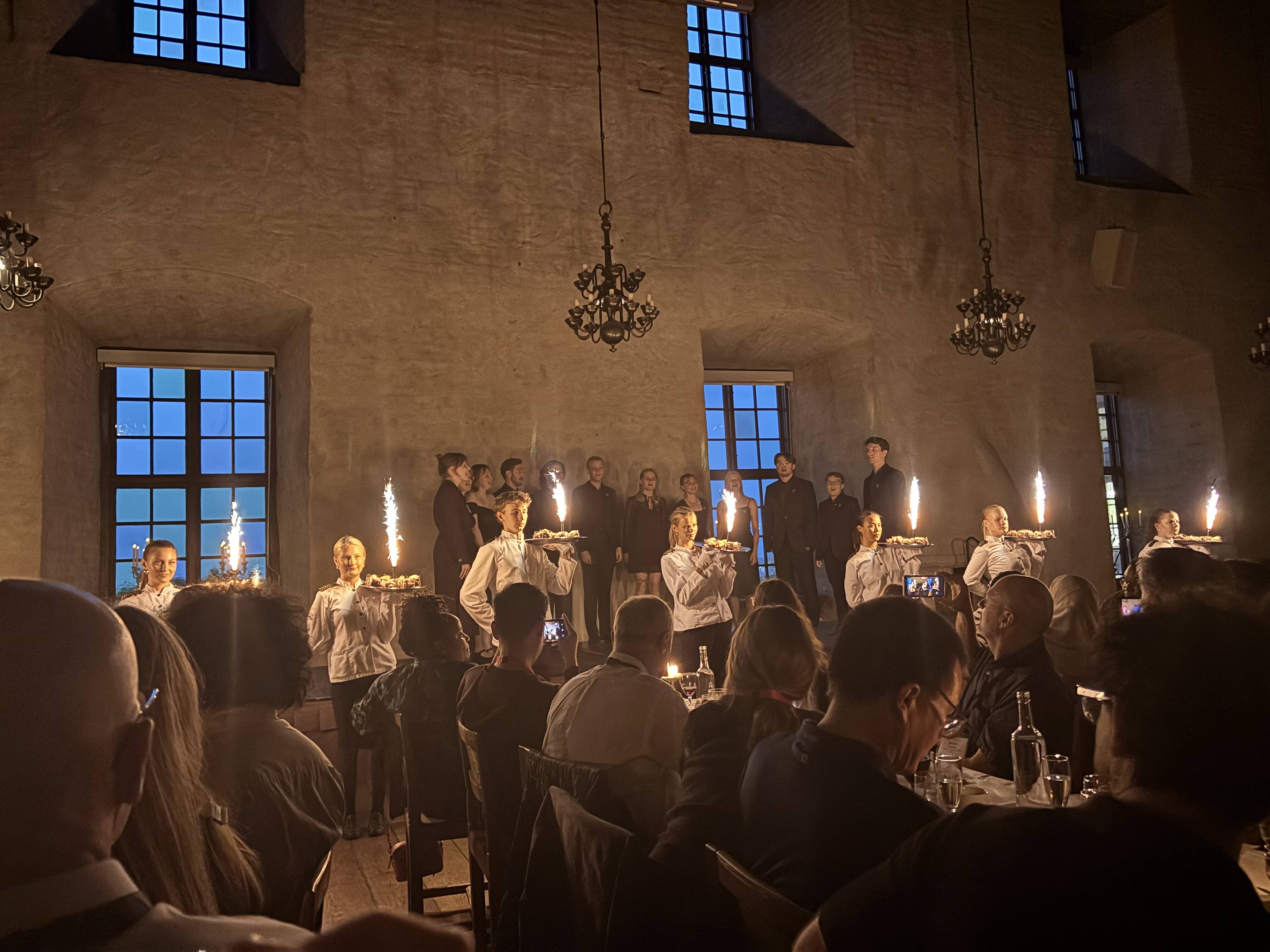 An impression from the banquet. On the stage is an a capella singing group, in front of them, waiters with desserts and small firework fountains, and in front of that, large tables with attendees sitting in long rows.